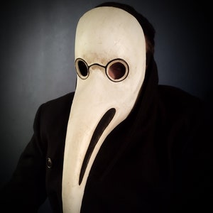 May include: A white plague doctor mask with a long beak and round eye holes. The mask has black accents around the eyes and beak.