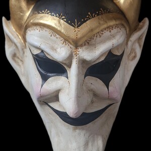 May include: A devil mask with a white face, black eyes and mouth, and gold horns. The mask has a black top with gold trim and is set against a black background. The mask has a sinister expression.