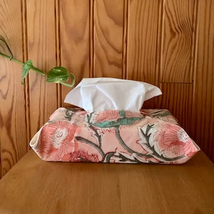 Peut inclure: Un couvre-boîte à mouchoirs en tissu floral rose avec un boîte à mouchoirs blanche à l'intérieur. Le couvre-boîte a un motif floral avec des couleurs rose, vert et blanc.