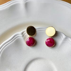 Peut inclure: Une paire de boucles d'oreilles dorées. Chaque boucle d'oreille présente un petit disque rond doré texturé en haut, relié à une pierre précieuse rose foncé, ronde et facettée, sertie dans une lunette dorée. Les boucles d'oreilles sont présentées sur une assiette blanche à festons.