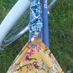 Könnte beinhalten: Ein blaues und weißes Blumenstoffband mit einem silbernen Metallclip, der an einem gelben Blumenstoff befestigt ist. Das Band ist an einem Fahrrad befestigt.