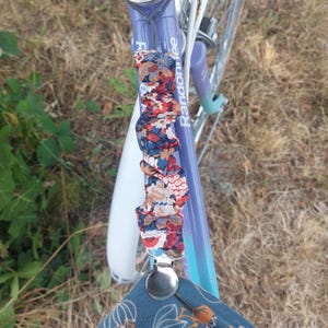 Op de afbeelding: Een close-up van een fietsstuur met een bloemenpatroon stoffen band en een zilveren clip. De band is bevestigd aan het stuur met een zilveren clip. De fiets is geschilderd in een lichtblauw en wit kleurenschema. De tekst "Randomfree" is zichtbaar op het stuur.