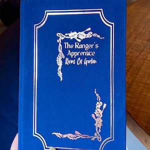 May include: A blue hardcover book with a silver border and a rose gold design. The title of the book is "The Ranger's Apprentice Ruins of Gorlan".