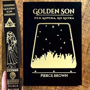 May include: A black hardback book with gold lettering titled "Golden Son: Per Aspera, Ad Astra" by Pierce Brown. The cover features a gold silhouette of a starry sky with a city skyline.