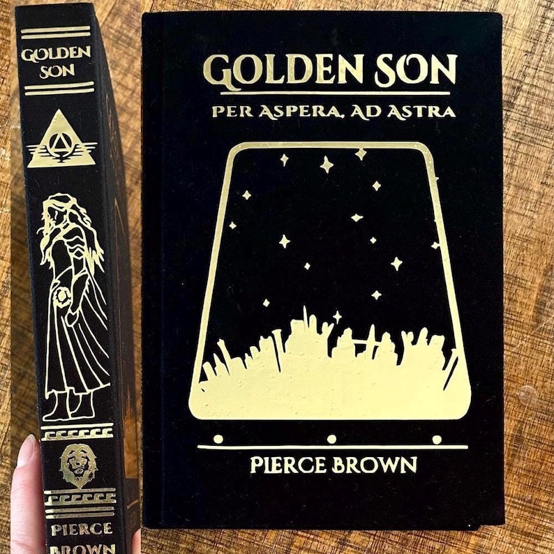 May include: A black hardback book with gold lettering titled "Golden Son: Per Aspera, Ad Astra" by Pierce Brown. The cover features a gold silhouette of a starry sky with a city skyline.