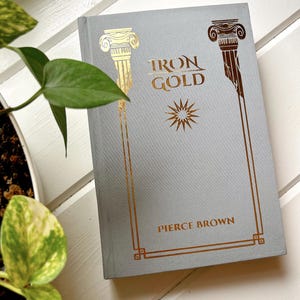 May include: A hardcover book titled "Iron Gold" by Pierce Brown. The book has a light gray cover with gold foil accents, including stylized columns and a starburst design. The book is resting on a white surface with a green plant in the corner.