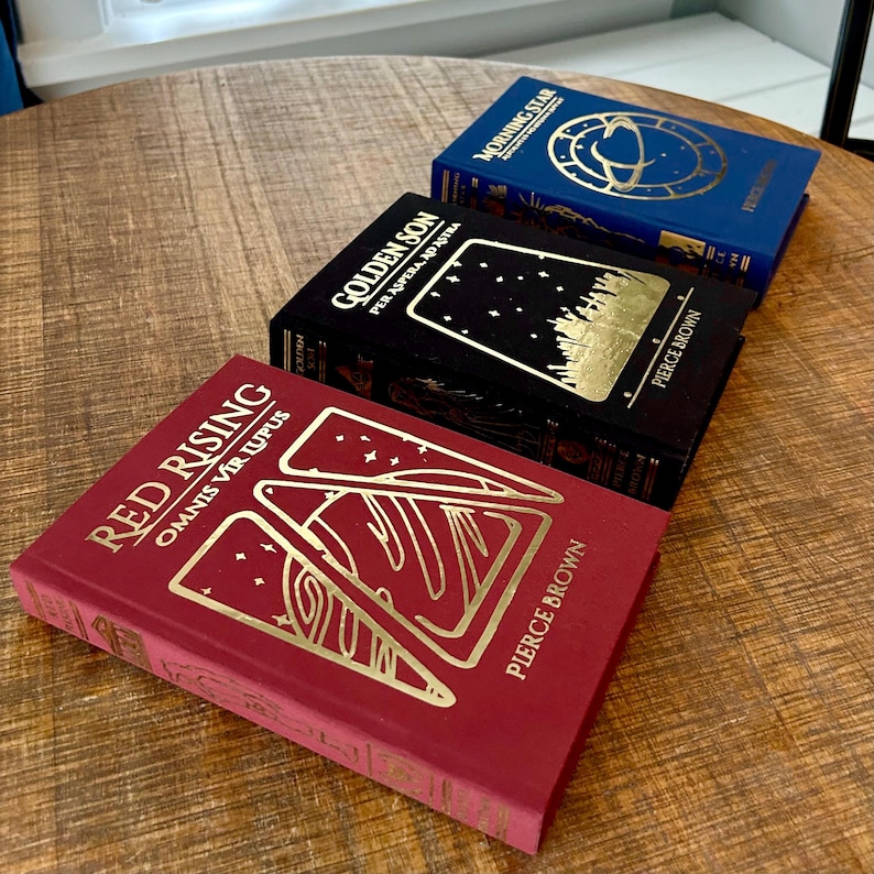 May include: Three hardback books stacked on a wooden table. The top book is blue with gold lettering and a gold illustration of a planet. The middle book is black with gold lettering and a gold illustration of a star field. The bottom book is red with gold lettering and a gold illustration of a mountain range.