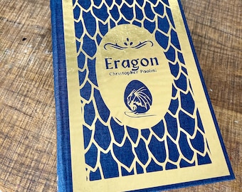 May include: A gold and blue hardcover book with the title "Eragon" by Christopher Paolini. The cover features a dragon egg design.