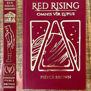 May include: A red hardback book with gold lettering titled "Red Rising" by Pierce Brown. The book cover features a stylised illustration of a planet with a gold outline.