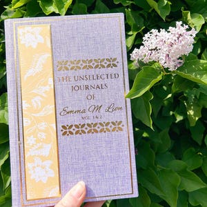 May include: A hardcover journal titled "The Unselected Journals of Emma M. Lion, Vol. 1 & 2." The journal has a lavender cover with gold accents and a yellow floral design on the spine. The background is green foliage and flowers.