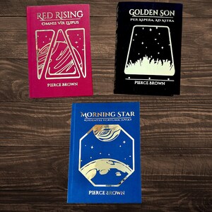 May include: Three hardback books with gold lettering and illustrations on the front covers. The books are titled "Red Rising", "Golden Son", and "Morning Star". The author is Pierce Brown.
