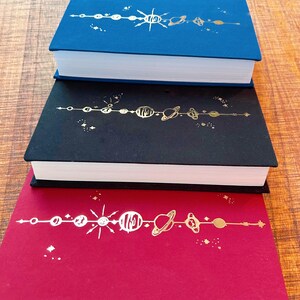 May include: Three hardback books with a gold solar system design on the spine. The top book is blue, the middle book is black, and the bottom book is red.