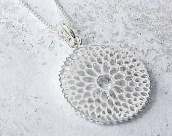 Può includere: Una collana con pendente in argento con un design floreale traforato e dettagliato. Il pendente circolare ha una pietra centrale e una catena sottile. La collana è mostrata su una superficie strutturata di colore chiaro.