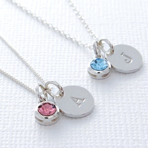 May include: Two silver necklaces with initial charms and small crystal accents. The necklace on the left has a pink crystal and the letter A charm. The necklace on the right has a blue crystal and the letter J charm.
