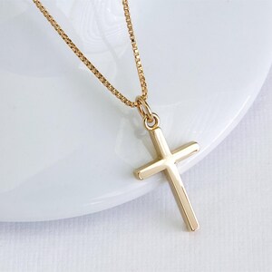 May include: A gold-colored cross pendant necklace. The cross is a simple, classic design with a polished finish. The chain is a delicate box chain style, also in gold. The necklace is displayed on a white surface.