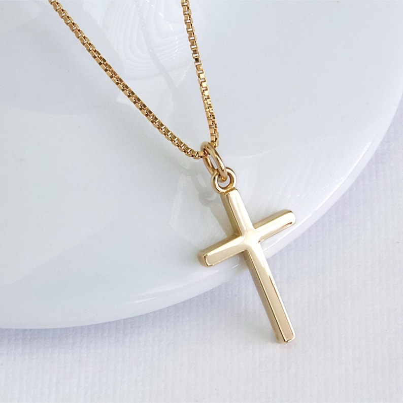 May include: A gold-colored cross pendant necklace. The cross is a simple, classic design with a polished finish. The chain is a delicate box chain style, also in gold. The necklace is displayed on a white surface.
