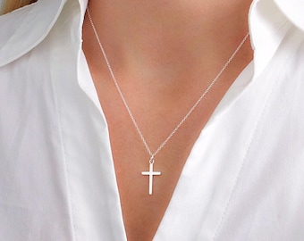 May include: A silver-toned metal cross pendant hangs from a delicate, thin silver-toned chain. The cross features a simple, smooth, and polished design with straight edges, suspended centrally against a crisp white collared shirt.