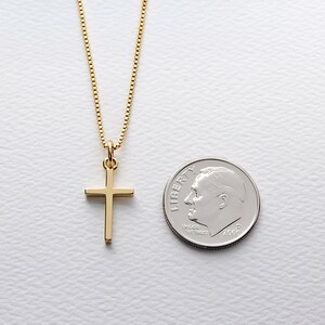 May include: A gold-colored cross pendant necklace. The cross is a simple, classic design, suspended from a delicate chain. A U.S. dime is next to the necklace for size comparison. The necklace is on a white surface.