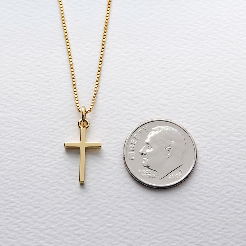 May include: A gold-colored cross pendant necklace. The cross is a simple, classic design, suspended from a delicate chain. A U.S. dime is next to the necklace for size comparison. The necklace is on a white surface.