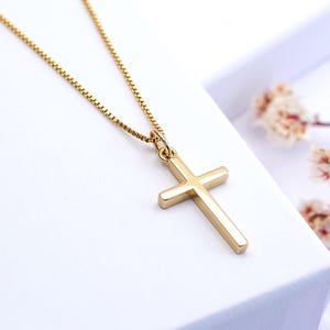 May include: A gold-colored cross pendant necklace. The cross is a simple, classic design with a polished finish. The chain is a delicate box chain style, also in gold. The necklace is displayed on a white surface.