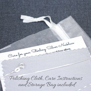 May include: A close-up of a silver necklace with a care instruction card and a polishing cloth. The card says "Care for your Sterling Silver Necklace" and "Polishing Cloth, Care Instructions and Storage Bag included".