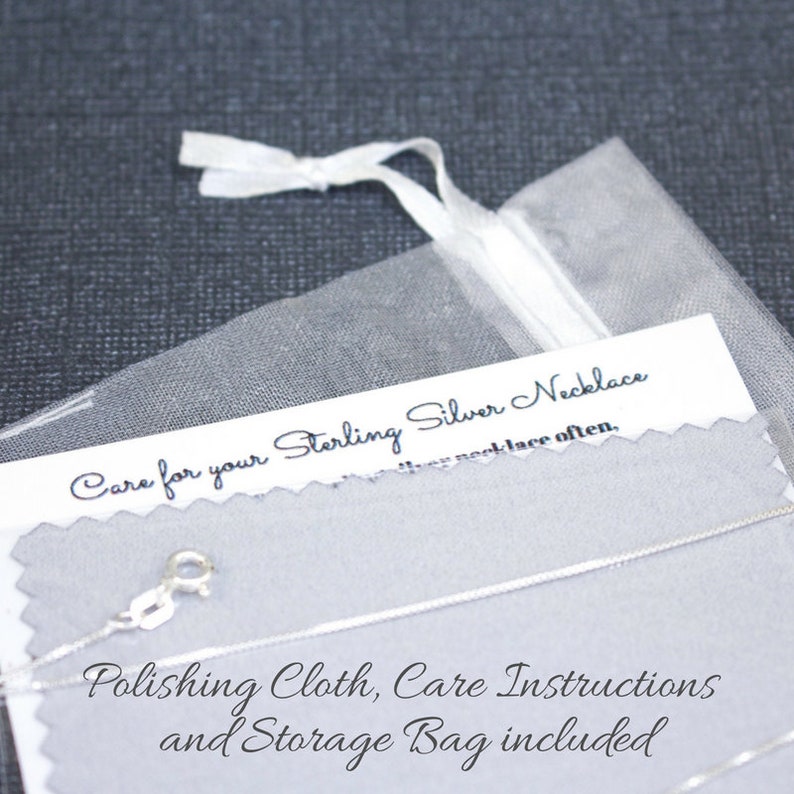 May include: A close-up of a silver necklace with a care instruction card and a polishing cloth. The card says "Care for your Sterling Silver Necklace" and "Polishing Cloth, Care Instructions and Storage Bag included".