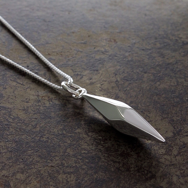 Puede incluir: Un collar de plata con un colgante geom&eacute;trico. El colgante tiene forma de diamante largo y puntiagudo y una superficie facetada.