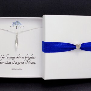 Puede incluir: Un collar de plata con un colgante geom&eacute;trico en una caja blanca con una cinta azul. La caja tiene el texto "No beauty shines brighter than that of a good Heart. 925 Sterling Silver."