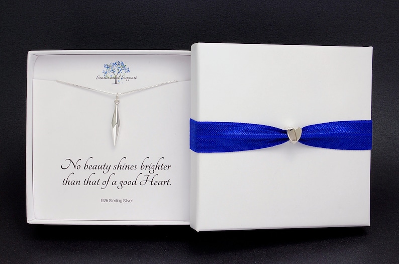 Puede incluir: Un collar de plata con un colgante geom&eacute;trico en una caja blanca con una cinta azul. La caja tiene el texto "No beauty shines brighter than that of a good Heart. 925 Sterling Silver."