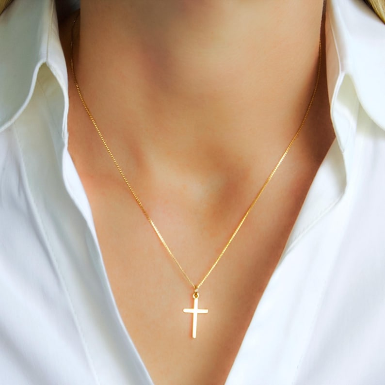 May include: A gold-colored cross pendant necklace. The cross is a simple, classic design, suspended from a delicate chain. The necklace is worn against a white shirt.