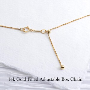 May include: Close-up of a 14k gold-filled adjustable box chain necklace. The chain is a bright gold color and features a delicate box link design. The adjustable portion of the chain has a small gold ball at the end. The text "14k Gold Filled Adjustable Box Chain" is at the bottom.