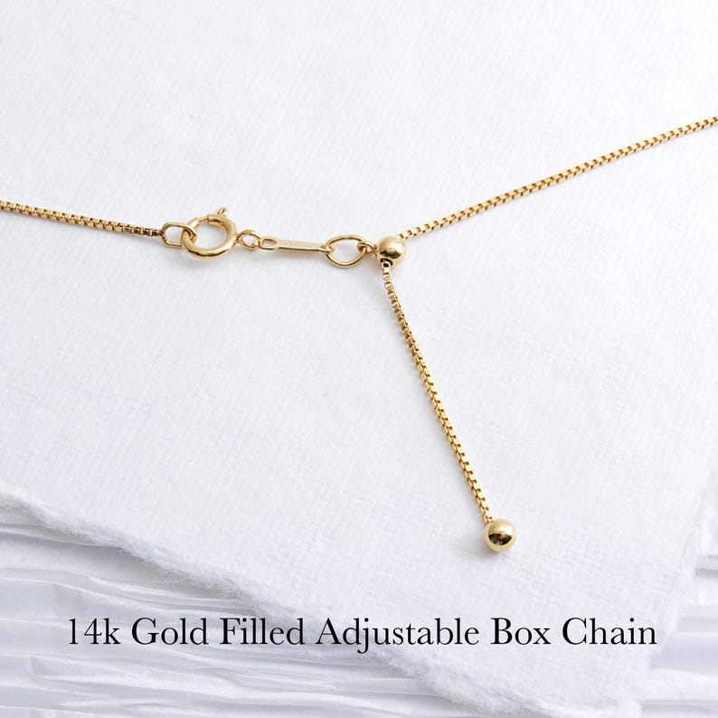 May include: Close-up of a 14k gold-filled adjustable box chain necklace. The chain is a bright gold color and features a delicate box link design. The adjustable portion of the chain has a small gold ball at the end. The text "14k Gold Filled Adjustable Box Chain" is at the bottom.