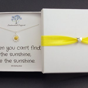 May include: A white gift box with a yellow ribbon tied around it. The box contains a silver necklace with a small daisy charm. The necklace is in a white box with the text "When you can't find the sunshine, be the sunshine. 925 Sterling Silver".