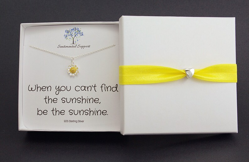 May include: A white gift box with a yellow ribbon tied around it. The box contains a silver necklace with a small daisy charm. The necklace is in a white box with the text "When you can't find the sunshine, be the sunshine. 925 Sterling Silver".