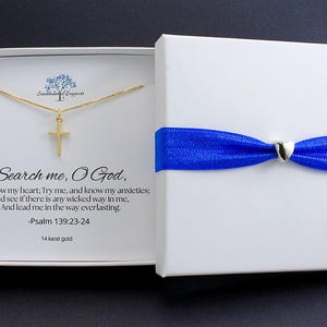 May include: A gold cross pendant necklace inside a white gift box. The box is tied with a blue ribbon and a silver heart-shaped clasp. The box's interior displays text from Psalm 139:23-24.