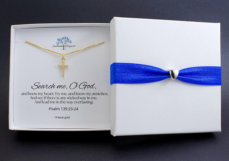May include: A gold cross pendant necklace inside a white gift box. The box is tied with a blue ribbon and a silver heart-shaped clasp. The box's interior displays text from Psalm 139:23-24.