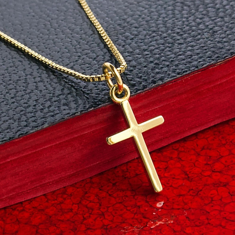 May include: A gold-colored cross pendant with a matching chain. The cross is simple in design and hangs from a delicate chain. The necklace rests on a dark blue and red surface, creating a contrast that highlights the jewelry.
