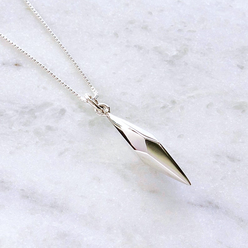 Puede incluir: Un collar de plata con un colgante geom&eacute;trico en forma de diamante. El colgante est&aacute; facetado y tiene una punta afilada.