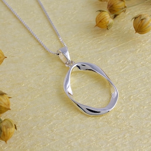 Könnte beinhalten: Eine silberfarbene Halskette mit einem Anhänger in Form eines fließenden, offenen Ovals. Der Anhänger hängt an einer filigranen Silberkette. Die Halskette wird auf einer hellgelben Oberfläche mit getrockneten floralen Akzenten präsentiert.