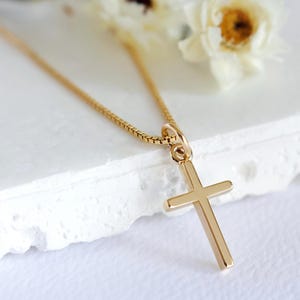 May include: A gold-colored cross pendant necklace. The cross is a simple, classic design with a rectangular shape. The chain is a delicate box chain. The background is a soft white surface with a few white flowers.