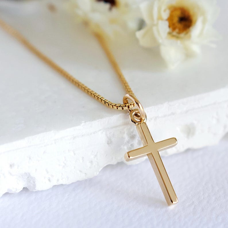 May include: A gold-colored cross pendant necklace. The cross is a simple, classic design with a rectangular shape. The chain is a delicate box chain. The background is a soft white surface with a few white flowers.