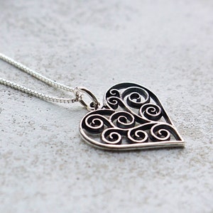 May include: A silver necklace with a delicate heart pendant featuring a detailed, intricate design of swirling scrolls.