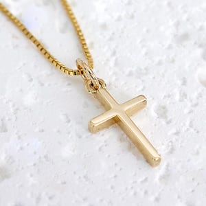 May include: A gold-colored cross pendant necklace. The cross is a simple, classic design with rounded edges. The chain is a delicate box chain style. The necklace is displayed on a textured white surface.