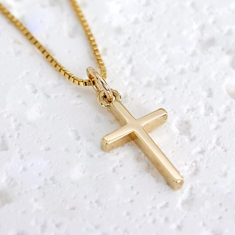 May include: A gold-colored cross pendant necklace. The cross is a simple, classic design with rounded edges. The chain is a delicate box chain style. The necklace is displayed on a textured white surface.