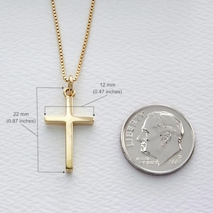 May include: A gold-colored cross pendant on a gold chain. The cross measures 0.87 inches wide and 0.47 inches high. A US dime is next to the cross for size comparison. The chain has a box link design.