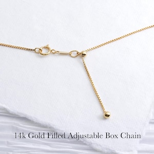 May include: Close-up of a 14k gold-filled adjustable box chain necklace. The chain is a bright gold color and features a delicate box link design. The adjustable portion of the chain has a small gold ball at the end. The text "14k Gold Filled Adjustable Box Chain" is at the bottom.