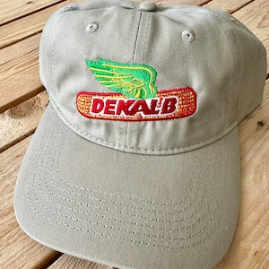 Op de afbeelding: Een kaki baseballpet met een geborduurd logo met een groene en gele vleugel met het woord "DENALB" in rode en gele letters.