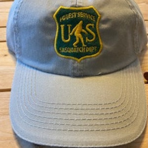 May include: A grey baseball cap with an embroidered patch featuring the text "Forest Service US Sasquatch Dept." in green and yellow.