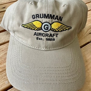 May include: A beige baseball cap with a black embroidered logo that says "Grumman Aircraft Est. 1929". The logo features a yellow winged design with a gray "G" in the center.