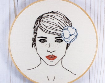 Pode incluir: Arte de bordado em aro que apresenta uma mulher com cabelo curto e liso e uma flor azul atrás da orelha. A mulher tem sardas e uma expressão neutra. O fundo é um tecido branco.